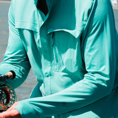 Person wearing a teal jacket holding a fly fishing reel by a body of water.
